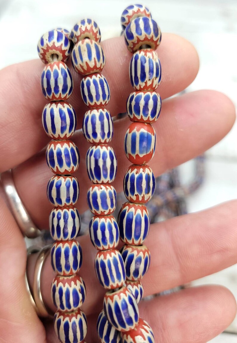May include: Strands of vintage beads with a repeating pattern. Each bead is a barrel shape with a red base, white stripes, and blue accents. The beads are strung together, creating a decorative element.