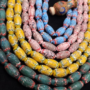 Op de afbeelding: Een ketting met kleurrijke, ovaalvormige kralen in tinten van teal, geel, roze en blauw. Elke kraal heeft een uniek patroon met rode en witte accenten. Een kleine, decoratieve vaas is ook aanwezig.
