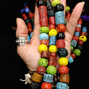 Puede incluir: Una colección de coloridos collares de cuentas con una variedad de formas y tamaños. Las cuentas son de color rojo, amarillo, verde, azul y negro. Los collares se sostienen en una mano sobre un fondo negro.