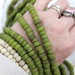 Puede incluir: Una hebra de cuentas de vidrio verde con una superficie texturizada. Las cuentas son redondas y tienen una forma ligeramente irregular. Las cuentas están enhebradas en un cordón y están listas para ser utilizadas para la elaboración de joyas.