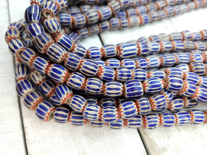 May include: Strands of vintage glass beads. The beads are oval-shaped with a white base, blue stripes, and red accents. The beads are strung together, creating a textured and colorful display. Ideal for jewelry making or crafting.