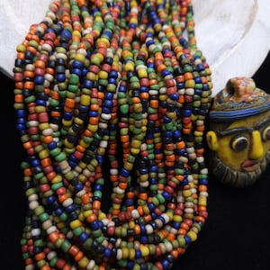 Könnte beinhalten: Eine Strähne bunter Glasperlen in den Farben Rot, Orange, Gelb, Grün, Blau, Schwarz und Weiß. Die Perlen sind klein und rund und auf einer durchsichtigen Schnur aufgereiht. Die Perlen sind in einem lockeren, fließenden Muster angeordnet.