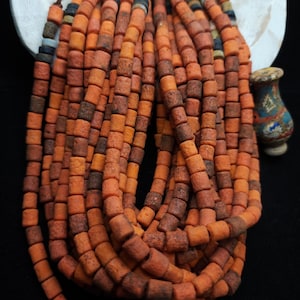 Può includere: Fili di perline quadrate arancioni e marroni. Le perline sono infilate insieme, creando un effetto strutturato e stratificato. Le perline hanno un aspetto rustico e terroso, che suggerisce un materiale naturale. La tavolozza dei colori è calda e invitante.