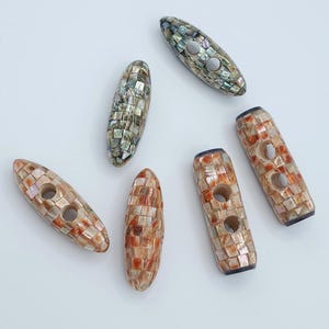 Puede incluir: Una colección de botones de palanca decorativos de varias formas y tamaños. Los botones están hechos de un mosaico de piezas de concha iridiscentes en tonos marrón, beige y gris. Cada botón tiene dos agujeros para coser.