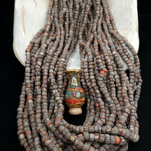 Puede incluir: Un collar de cuentas a rayas grises y naranjas, con un pequeño jarrón decorativo en el centro. El jarrón está pintado con diseños florales coloridos. Las cuentas son cilíndricas y están dispuestas en múltiples hebras.