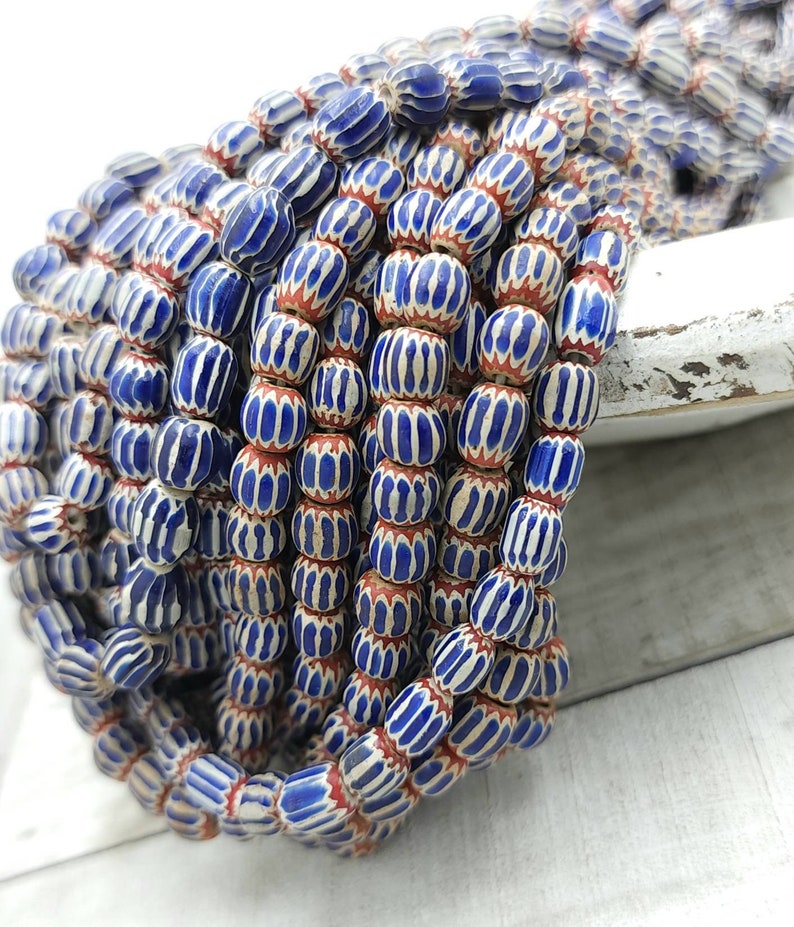 May include: A collection of blue and white striped beads with red accents. The beads are oval-shaped and strung together, creating a textured and colorful display. The beads are likely for jewelry making.