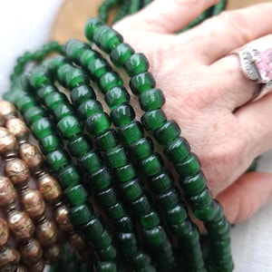 Puede incluir: Primer plano de una hebra de cuentas de vidrio verde oscuro. Las cuentas son redondas y tienen una forma ligeramente irregular. Están enhebradas en un hilo transparente.