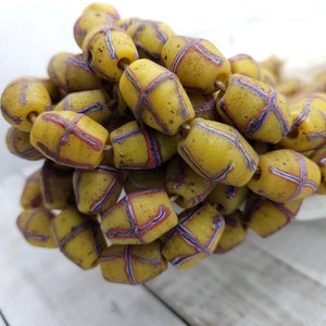 May include: A collection of yellow, barrel-shaped beads, each adorned with a red and blue cross-hatch pattern. The beads have a slightly worn look. They are strung together, suitable for jewellery making or other craft projects. Each bead is approximately 1.5 cm in length.