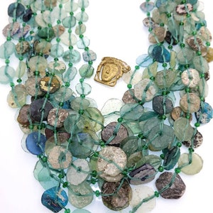 May include: A multi-strand necklace made of green and blue glass beads. The beads are of various shapes and sizes, and some have a weathered look. A small bronze pendant with a face is in the center of the necklace.