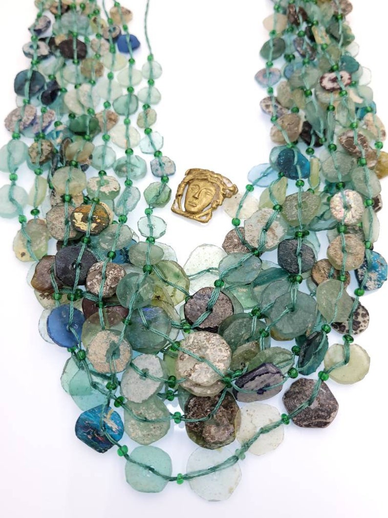 May include: A multi-strand necklace made of green and blue glass beads. The beads are of various shapes and sizes, and some have a weathered look. A small bronze pendant with a face is in the center of the necklace.