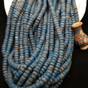 Pode incluir: Um fio de contas de vidro azul com uma superfície áspera e texturizada. As contas são redondas e têm uma forma ligeiramente irregular.