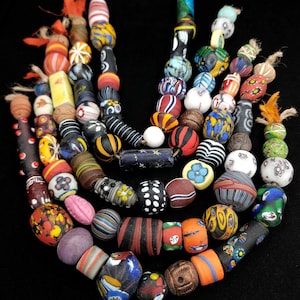 May include: A collection of multi-colored glass beads arranged in several strands. The beads feature various patterns including stripes, dots, floral motifs, and solid colors in shades of red, blue, yellow, green, black, and white, with frayed fiber ends.