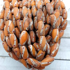 Puede incluir: Un montón de cuentas de vidrio naranja y blanco con acentos rojos. Las cuentas son de forma ovalada y tienen una superficie texturizada.