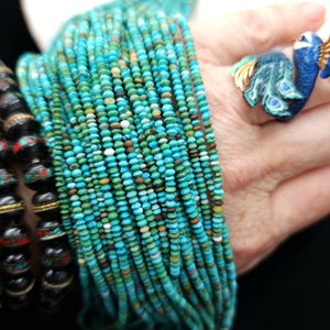 May include: A strand of turquoise beads, with a variety of shades of blue and green, is wrapped around a person's wrist. The beads are small and round, and they are strung on a thin wire.