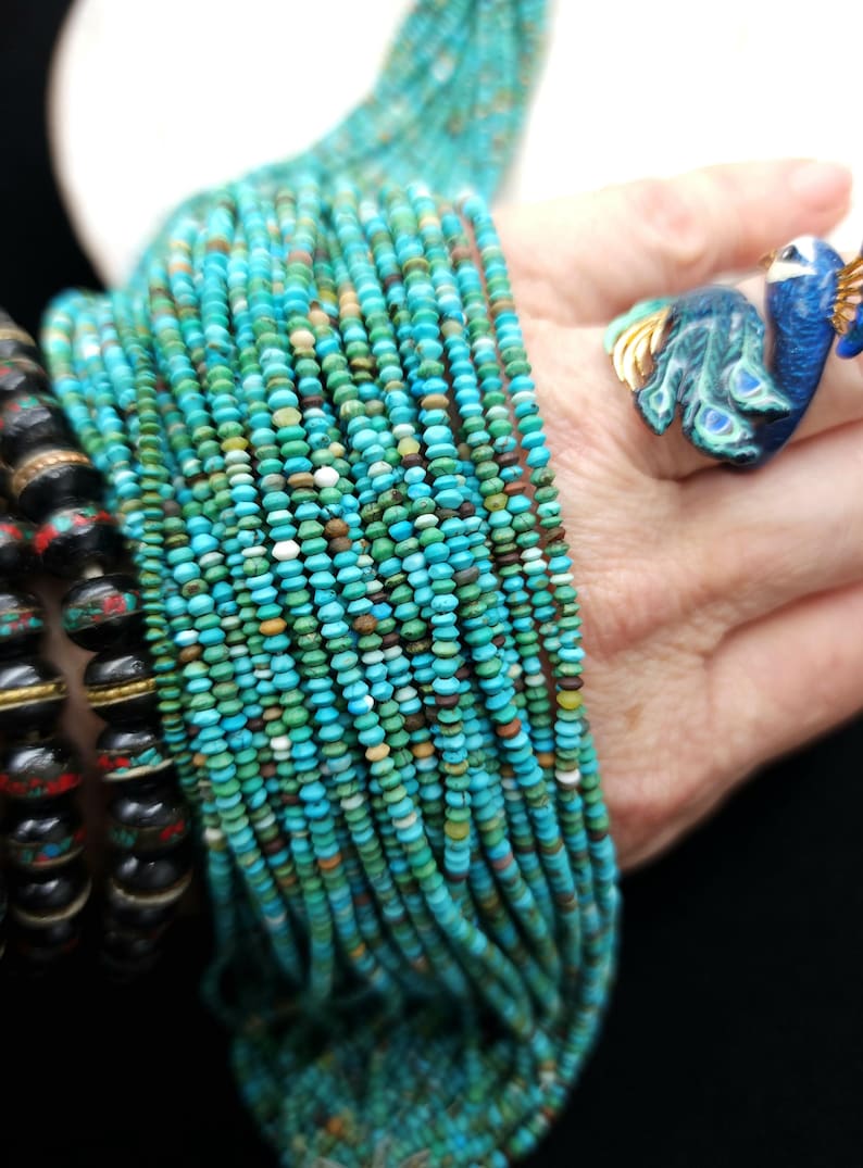 May include: A strand of turquoise beads, with a variety of shades of blue and green, is wrapped around a person's wrist. The beads are small and round, and they are strung on a thin wire.