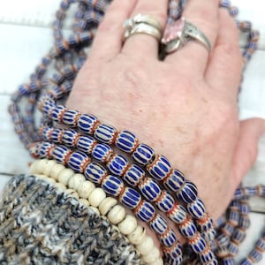 May include: Strands of vintage glass beads in blue, white, and red. The beads have a striped pattern and are oval-shaped. A few strands of off-white, round beads are also visible. The beads are displayed on a hand with rings.