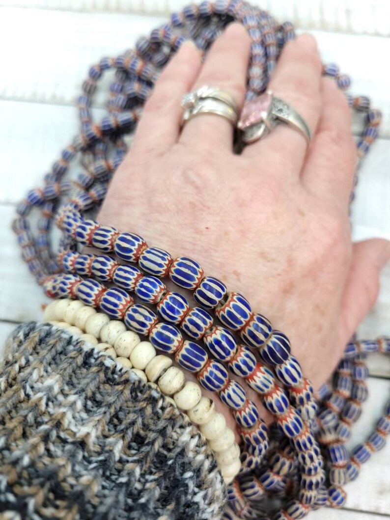 May include: Strands of vintage glass beads in blue, white, and red. The beads have a striped pattern and are oval-shaped. A few strands of off-white, round beads are also visible. The beads are displayed on a hand with rings.