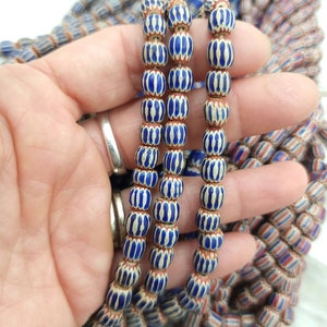 May include: Strands of blue and white striped beads with a brown base are held in a hand. The beads have a barrel shape and are strung together. Other strands of beads with red, white, and blue stripes are visible in the background.