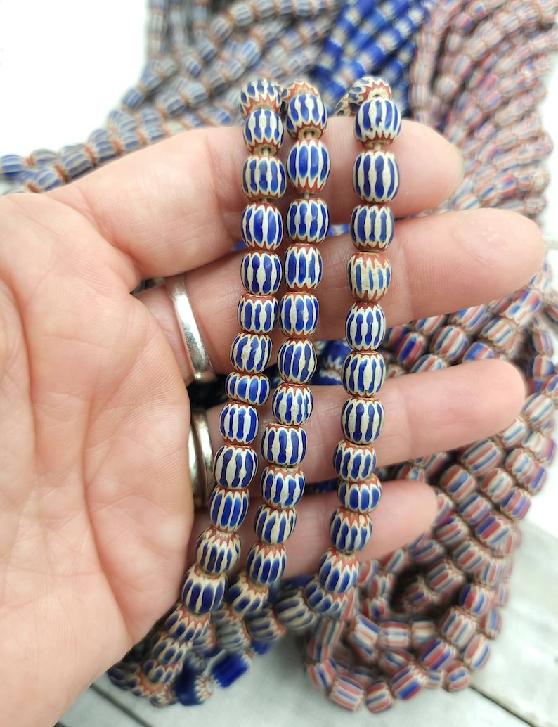 May include: Strands of blue and white striped beads with a brown base are held in a hand. The beads have a barrel shape and are strung together. Other strands of beads with red, white, and blue stripes are visible in the background.