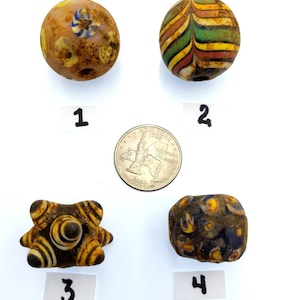 May include: Four antique glass beads with various colors and patterns. The beads are numbered 1 through 4. Bead 1 is a round bead with a light brown background and blue, white, and yellow accents. Bead 2 is a round bead with a white background and yellow, green, red, and brown stripes. Bead 3 is a star-shaped bead with a black background and yellow and white stripes. Bead 4 is a round bead with a dark blue background and yellow, white, and red accents.