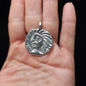 May include: Silver pendant featuring a detailed profile of a woman's face with a crown and intricate hair. The pendant is circular and has a loop at the top for attaching a chain.