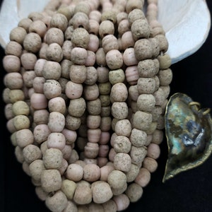 Pode incluir: Um fio de contas de cor rosa claro e bege feitas de pedra natural. As contas são redondas e têm uma superfície áspera e texturizada. O fio é longo e está drapeado sobre uma superfície branca.