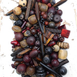 以下が含まれることがあります： 茶色、黒、赤、自然な木の色合いなど、さまざまな形、サイズ、色の木製ビーズのコレクション。ビーズは白い表面のボウルに配置されています。