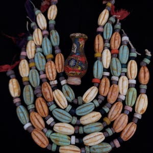 Pode incluir: Um colar com vários fios de contas ovais em tons de creme, laranja, azul e verde. Um pequeno vaso colorido está centrado e o colar tem borlas decorativas.