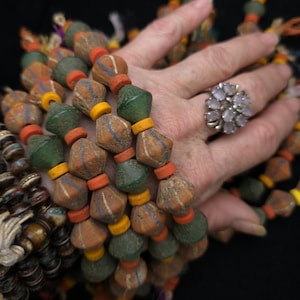 May include: Close-up of colourful beaded necklaces. The beads are a mix of orange, green, and yellow, with intricate patterns. A silver ring with a cluster of light purple stones is also visible. The necklaces are likely handmade.