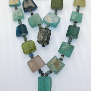 May include: A necklace made of various colored glass beads. The beads are square and rectangular in shape and are strung on a cord. The necklace has a gold pendant in the shape of a head.