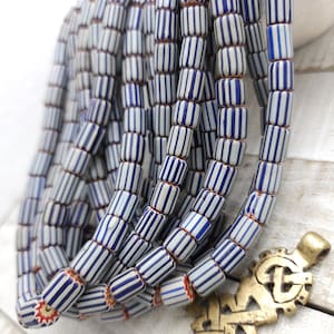 Puede incluir: Una hebra de cuentas de vidrio rayadas en azul y blanco. Las cuentas son cilíndricas y tienen una forma ligeramente irregular. Las cuentas están enhebradas en un hilo transparente.