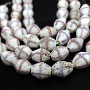 Puede incluir: Una hebra de cuentas de vidrio blanco con líneas azules y rojas. Las cuentas son facetadas y tienen una forma ligeramente irregular. Las cuentas están enhebradas en un cordón delgado.