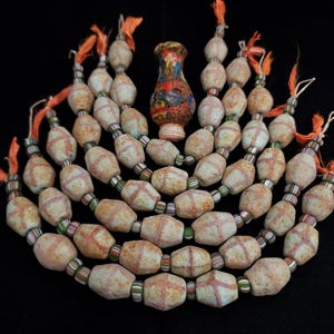 Puede incluir: Una colección de cuentas antiguas y un pequeño jarrón sobre un fondo negro. Las cuentas blanquecinas presentan diseños rojos de trama cruzada. El jarrón es multicolor con una base redondeada y un cuello estrecho. Borlas naranjas están unidas a los hilos de cuentas.