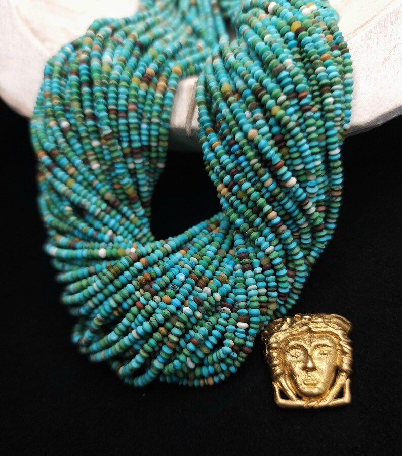 May include: A long strand of turquoise beads with a gold pendant featuring a stylized human face. The beads are a mix of blue, green, and brown colors.