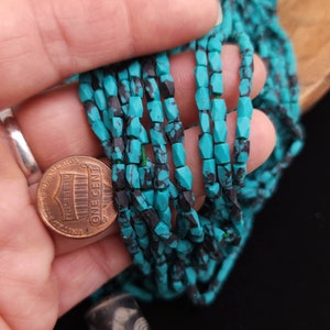 May include: A strand of turquoise beads with black matrix. The beads are faceted and have a rectangular shape. The beads are strung on a clear thread. A US penny is included for scale.