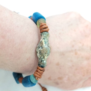 Puede incluir: Un brazalete de cuero con cuentas de vidrio azul y verde y una gran cuenta de metal desgastada en el centro.