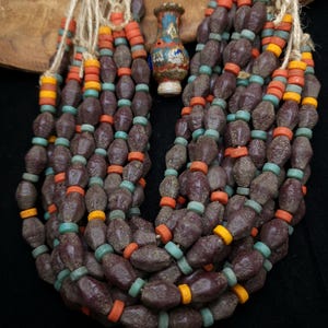 以下が含まれることがあります： 表面にテクスチャのあるダークブラウンの楕円形のビーズでできたマルチストランドネックレス。ネックレスは、オレンジ、ターコイズ、イエローの小さな丸いビーズでアクセントが付けられています。