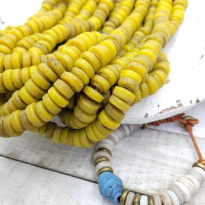 Puede incluir: Una hebra de cuentas de vidrio amarillo con una hebra más pequeña de cuentas de vidrio blanco y azul. Las cuentas son todas redondas y tienen una textura ligeramente rugosa.