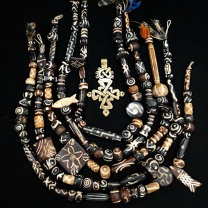 May include: A collection of multiple strands of dark brown and cream-colored patterned beads, featuring various shapes including cylinders, spheres, squares, and fish-shaped pendants, centered around a single ornate brass cross pendant.