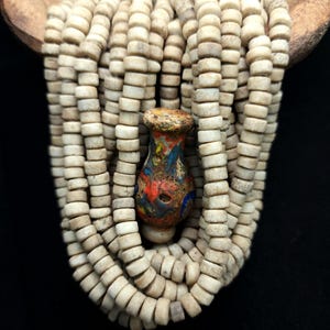 May include: A strand of off-white bone beads with a single, colorful, glass bead in the center. The glass bead is a mottled blue, red, and white with a gold-colored top.