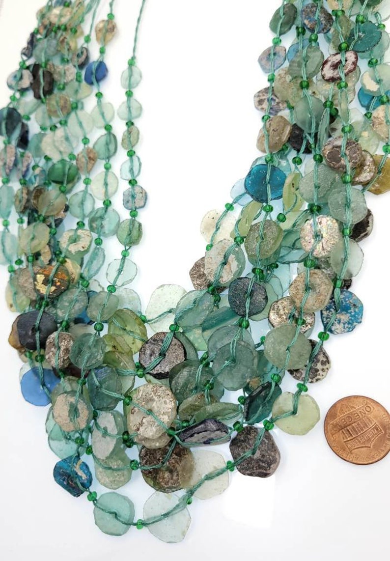May include: A multi-strand necklace made of green, blue, and clear glass beads. The beads are various shapes and sizes, and some have a weathered look. The necklace is about 18 inches long and is shown next to a US penny for scale.