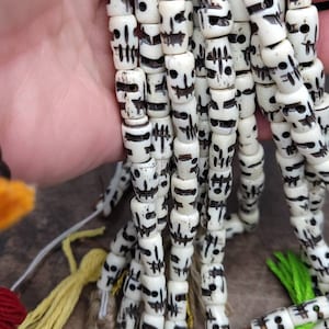 May include: A strand of white skull beads with black painted details. The beads are strung on a white cord and are approximately 1 cm in diameter.
