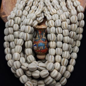 Pode incluir: Colar de contas creme com um pequeno pingente ornamental em forma de vaso. As contas têm uma textura canelada e são enfiadas. O pingente é multicolorido com detalhes azuis, vermelhos e dourados. O colar é exibido em um fundo escuro.