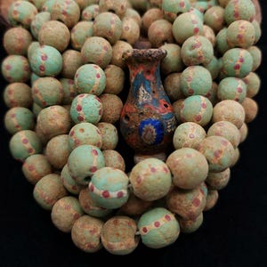 May include: A collection of round, textured beads in shades of light green and tan, each featuring a central band with small red dots. A small, ornate, multi-coloured vase-shaped pendant with blue, red, and white patterns sits in the centre.