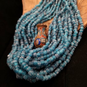 以下が含まれることがあります： 複数のストランドに配置された、ターコイズ色のガラスビーズのコレクションと、小さな装飾的な花瓶。花瓶は主に赤と茶色で、青と白のアクセントがあります。ビーズはテクスチャード加工されています。