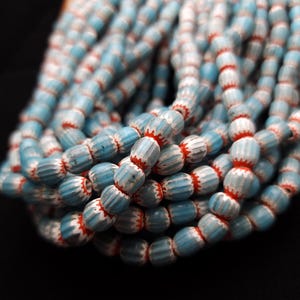 Pode incluir: Um fio de contas de vidro azul e branco com detalhes vermelhos. As contas são cilíndricas e têm uma textura estriada.