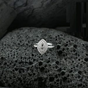 May include: A silver ring with a marquise-shaped cluster of small, sparkling stones. The ring's band is thin and delicate, and the setting is a bright, reflective silver. The ring is set against a dark, textured background.