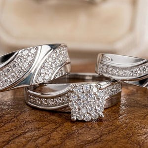 Puede incluir: Un conjunto de tres anillos plateados con brillantes piedras preciosas transparentes. Los anillos se exhiben sobre una superficie marrón. El anillo central presenta un grupo cuadrado de piedras, los otros tienen un diseño ondulado con piedras incrustadas.