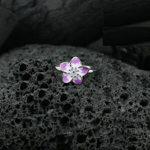 May include: A silver ring featuring a flower design with purple and white enamel petals. The flower has a small, white center. The ring is set against a dark, textured background.