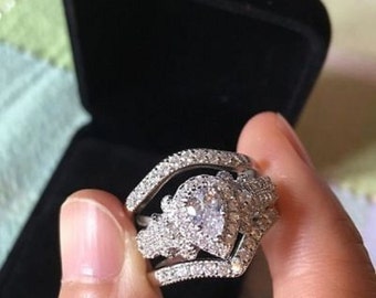 Puede incluir: Un anillo de plata con un gran diamante de forma de pera rodeado de diamantes más pequeños. El anillo está engastado en una banda con un diseño delicado.